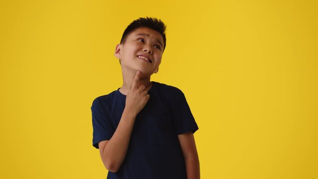 Young asian boy puts his finger to his chin and thinks hard with an exhausted grimace on his face on a yellow background. Concept of a difficult choice of useful activity for a child