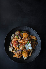 Fusilli pasta with mushrooms, asparagus and cherry tomatoes on a modern design plate with dark background