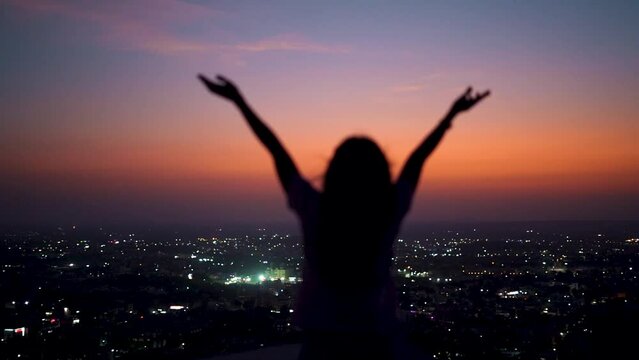 Silhouette of defocused Indian girl raises her hands up in front of city during night. Travel concept. Concept of welcoming, relaxing and enjoying.