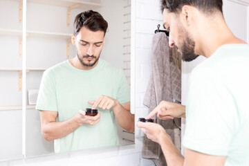 Handsome man looking at mirror and applying moisturizing cream on cheeks in bathroom. Groomed young guy doing skincare morning routine after taking a shower