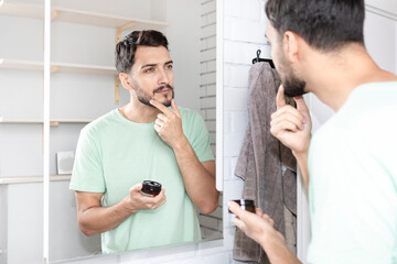 Handsome man looking at mirror and applying moisturizing cream on cheeks in bathroom. Groomed young guy doing skincare morning routine after taking a shower