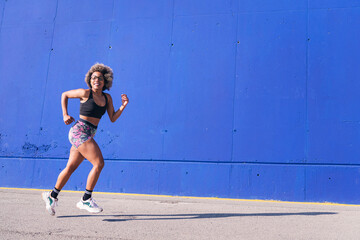 african american woman running in an urban track with blue background, concept of sport and active lifestyle, copy space for text