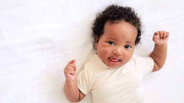 little african American baby girl in a white bodysuit on a cotton bed at home, six-month-old smiling joyful black baby