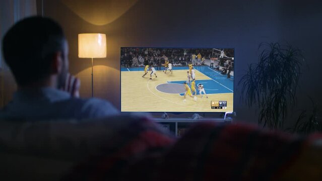 Back View of a Middle Aged Man Sitting on a Sofa at Home in the Evening and Watching a Live Basketball Broadcast on a Big Flat Screen TV. Male Relaxing at Home, Cheering For His Favorite Team