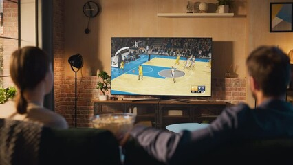 Young Couple Spending Time at Home, Sitting on a Couch and Watching and Exciting Live Basketball Tournament on TV in Their Stylish Loft Apartment. Man and Woman Eating Snacks and Enjoy the Sports Show