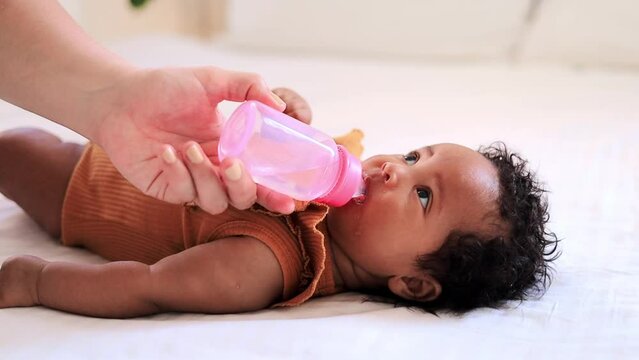 a small African-American baby girl drinks water or milk from a bottle in an orange bodysuit on a white bed, her mother's hand feeds a six-month-old black newborn baby from a bottle