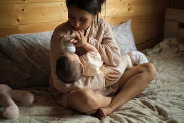 Sitting on the bed, a young mother feeds her baby with a specialized formula, demonstrating the diverse choices parents make today, sometimes opting for or relying on alternative feeding methods.
