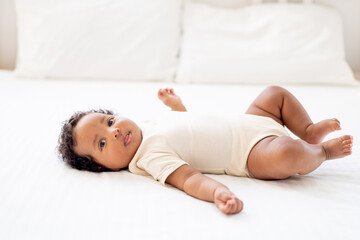 Obraz na płótnie Canvas a close-up portrait of a small African-American baby girl in a white bodysuit on a cotton bed at home, a funny six-month-old smiling joyful black newborn baby lies on the back