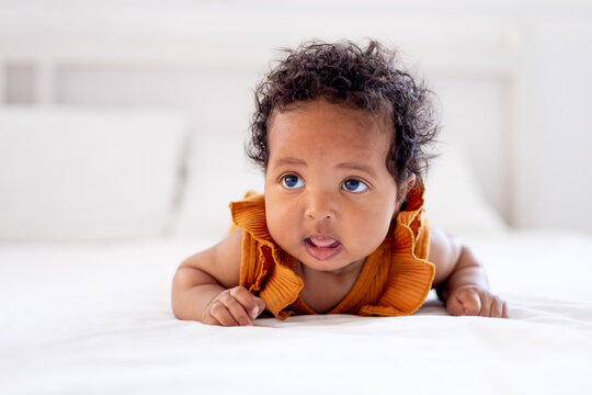funny little African-American baby girl in an orange bodysuit on the bed at home shows her tongue, funny six-month-old black newborn baby lies on her tummy and rejoices. View from above