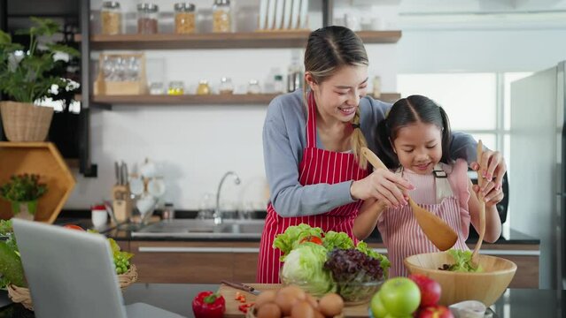 Beautiful young asian mother teaching daughter making vegetable salad in kitchen room at home. Happy mother and daughter in apron enjoy cooking together. Family Activity, Healthy food concept