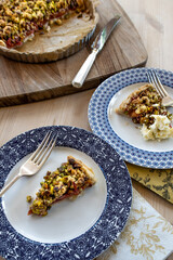 Slices of plum pie with a pistachio crumble topping served on attractive plates with clotted cream on one