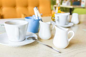 Breakfast service. Coffee, milk and sugar in white utensil. Coffee mug and milk jug on the table in cafe. Cafe interior. White tableware. Morning coffee concept. 