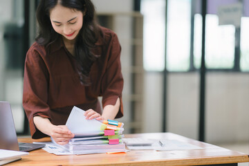 The businesswoman's hands are busy working amidst stacks of paper files, searching and checking for...