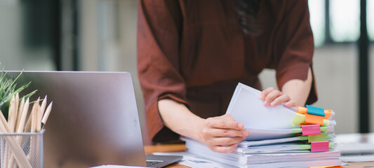 The businesswoman's hands are busy working amidst stacks of paper files, searching and checking for...