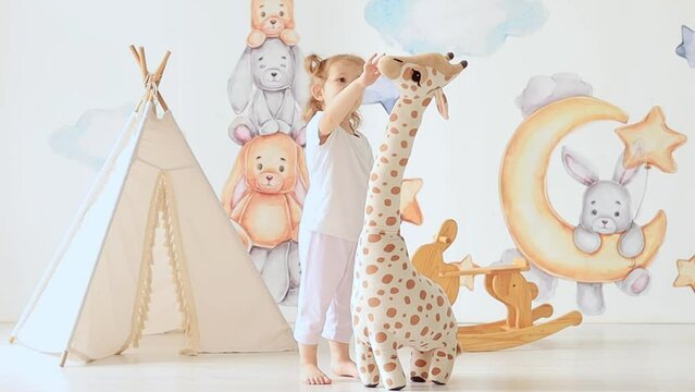 little child girl blonde plays in the children's room, hugs a big toy giraffe, measures her height, children's leisure, kindergarten