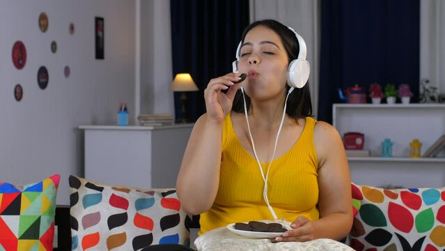 Women relaxing at home - eating cookies and listening to music on headphones  high sugar content  unhealthy lifestyle  binge eating . Young Indian girl enjoying a plate full of biscuits - unhealthy...