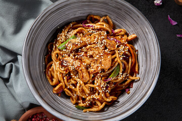 Overhead, horizontal view of stir-fried udon noodles with chicken and vegetables, beautifully arranged on a black background, with spices and a napkin lending character