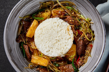 Top view of Chinese stir fry with rice and meat, combined with pineapples in a sauce in a gray bowl. Arrangement includes a black background, fabric, and chili peppers