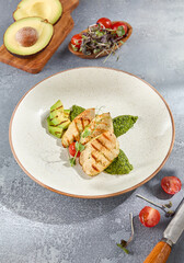 Grilled chicken fillet paired with avocado and spinach on a ceramic plate. The composition is set against a grey concrete background with hard shadows, making it ideal for healthy meal concepts