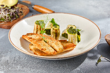 Closeup side view of grilled perch with zucchini rolls on a ceramic plate. The hard shadows on the gray concrete background add a dramatic touch. Perfect for a gourmet menu