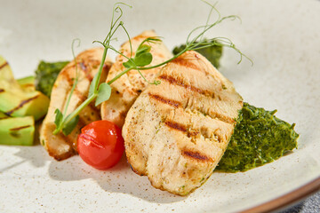 A macro side view of grilled chicken fillet with avocado and spinach on a ceramic plate. The intricate textures come alive against a grey concrete background with hard shadows