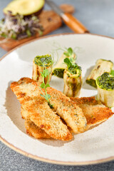 Vertical closeup side view of grilled perch and zucchini rolls on a ceramic plate. The dish is highlighted by the gray concrete background and hard shadows