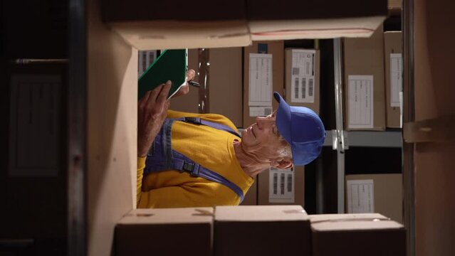 Elderly warehouse worker preparing goods for shipment while standing with clipboard near shelves with parcels ready for shipment.