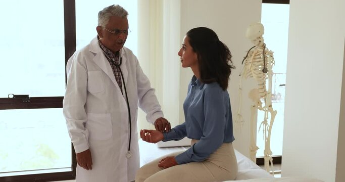 Senior male Indian doctor in uniform measure heart rate manually by hand placing fingers on female patient wrist counts number of beats. Young woman sit on medical couch while GP check her heartbeat