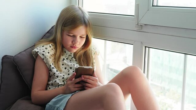 Cute teenage girl with a smartphone sitting on the windowsill and looking down. 