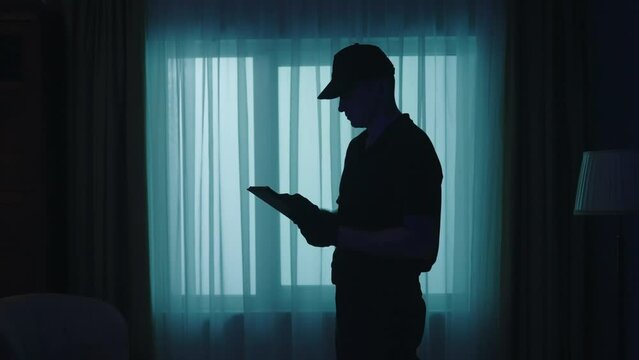 Closeup shot of policeman in uniform with tablet standing in the dark living room, looking around, writing something. Crime scene creative concept.