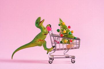 Happy green toy dinosaur carrying shopping cart with Christmas tree on pastel pink background....