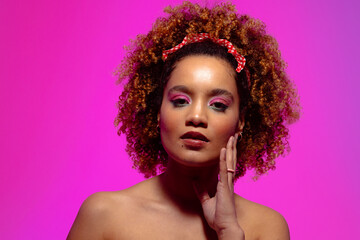 Biracial woman with curly hair, pink eye shadow and lipstick touching face on pink background