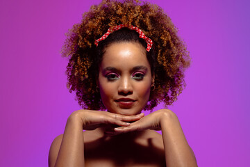 Biracial woman with curly hair, pink eye shadow and lipstick, hands under chin on purple background