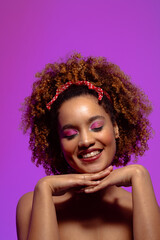Smiling biracial woman with pink eye shadow, eyes closed and hands under chin, purple copy space