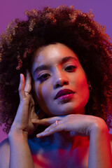 Biracial woman with curly hair, touching face in red and blue light with purple background