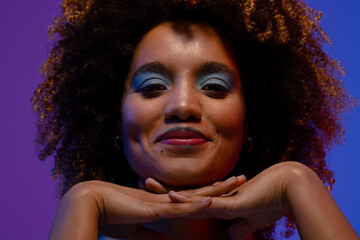 Smiling biracial woman with curly hair and blue eye shadow with hands under chin