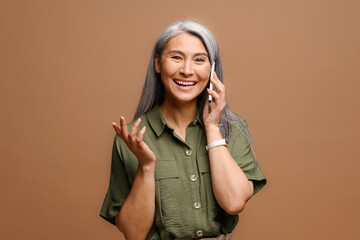 Cheerful happy mature Asian woman talking on smartphone isolated on brown background, 40s lady...
