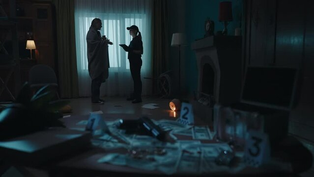 Young woman victim in blanket talking with police officer in the dark apartment room with signs of murder and robbery. Crime scene creative concept.