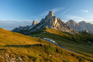 Fotobehang Dolomieten Sunrise at Passo Giau with road and parked cars and campers with sun shining at meadow and peaks in  the background during summer morning in august and grass in foreground