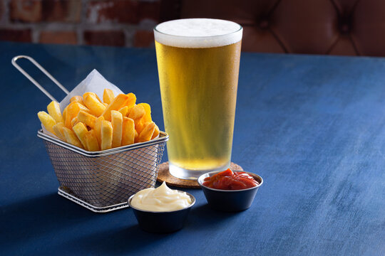 Golden French Fries in a Wire Basket with a Pilsner or Lager Beer and Ketchup and Mayonnaise Dipping Sauces in a Bar