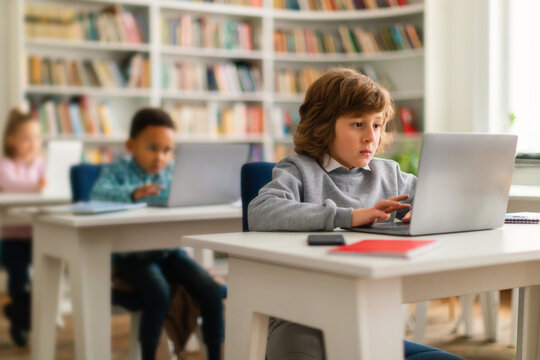Computer science class. School boy sitting at desk at classroom with classmates on background, typing on laptops