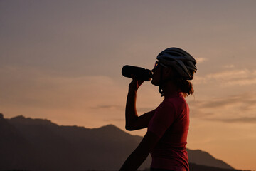 Cyclist silhouette. Female cyclist in cycling clothes and a helmet drinks water from a sports bottle at sunset.