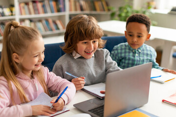 Three diverse classmates sitting in front of laptop, watching videos online during lesson, looking at screen and smiling