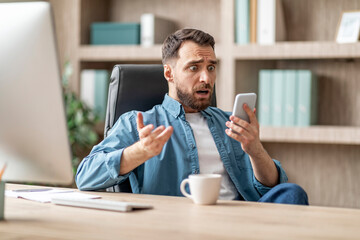 Shocked Businessman Reading Message On Smartphone While Sitting At Workplace In Office
