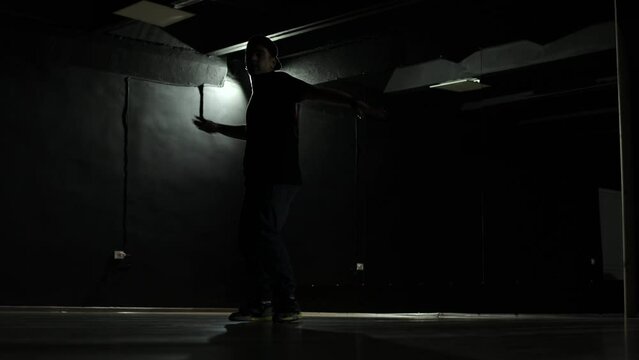 A man is dancing hip-hop, his silhouette is visible in the dark. A dark hall, one light bulb, a boy dancing. The man likes hip-hop, dancing in the dark. At night, a person dances.