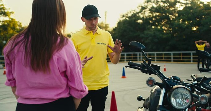 A driving instructor in a yellow t-shirt tells a female student in a pink t-shirt how to behave while driving a motorcycle