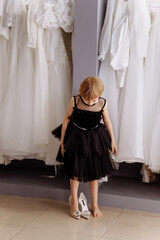 Beautiful white wedding dresses are hanging on hangers in the bridal salon. A little girl chooses and tries on a black dress and oversized shoes.