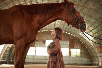 Side view. Cute little girl is with horse indoors