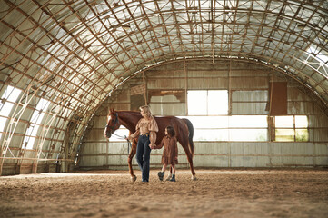 Distant view. Woman with her little daughter is with horse indoors