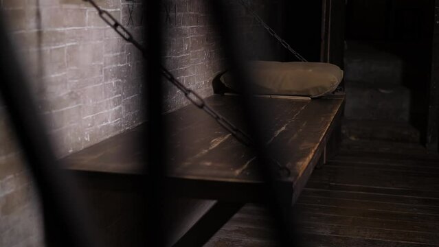 A close-up of a wooden bed hanging on chains, a shelf with a pillow in an old prison cell. A dark room with a solid wooden bed hanging on chains. An old prison.